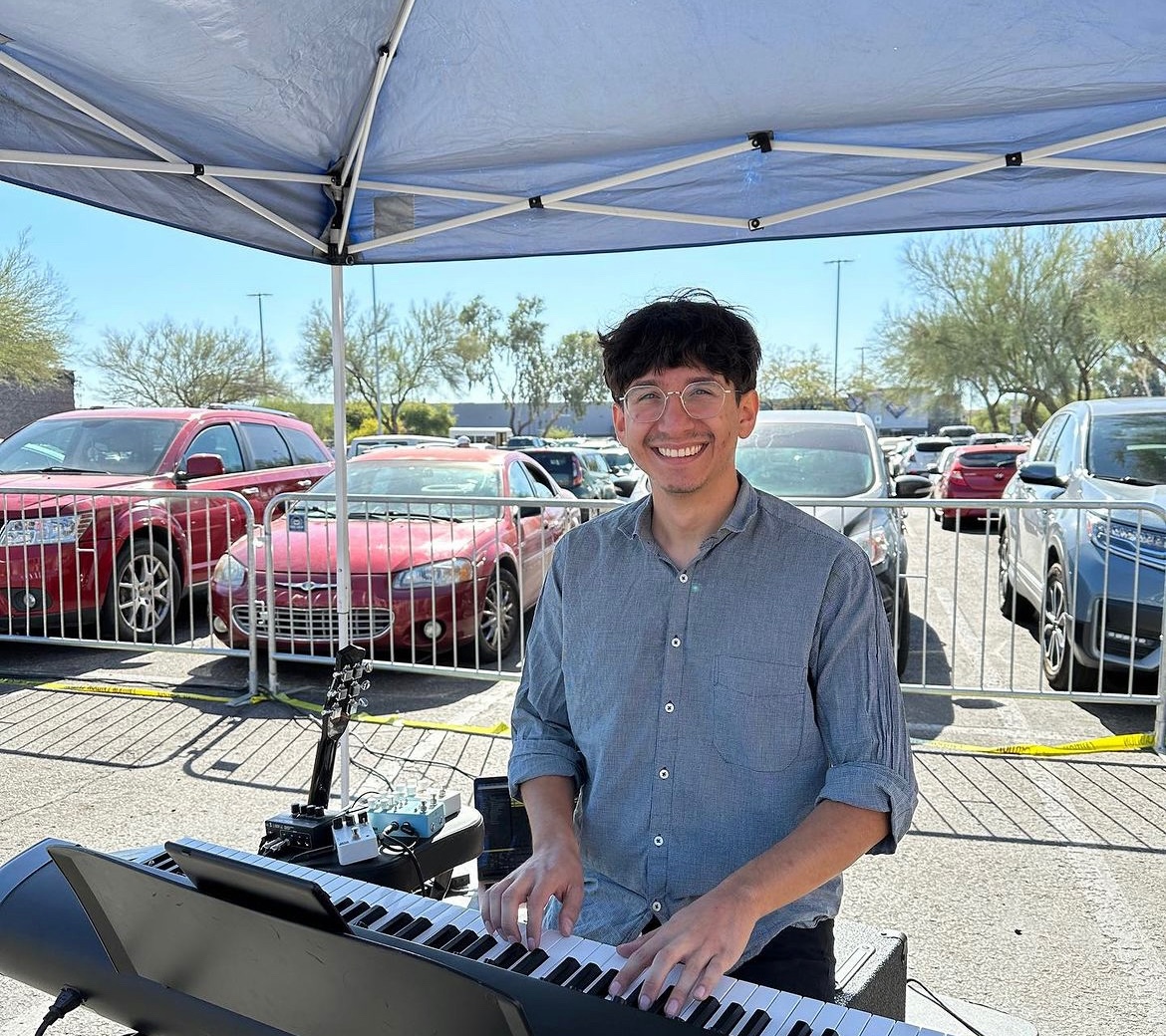 Daniel seated at a keyboard at an outdoor event, smiling at the camera. He has brown hair and blue eyes and is wearing a blue shirt.